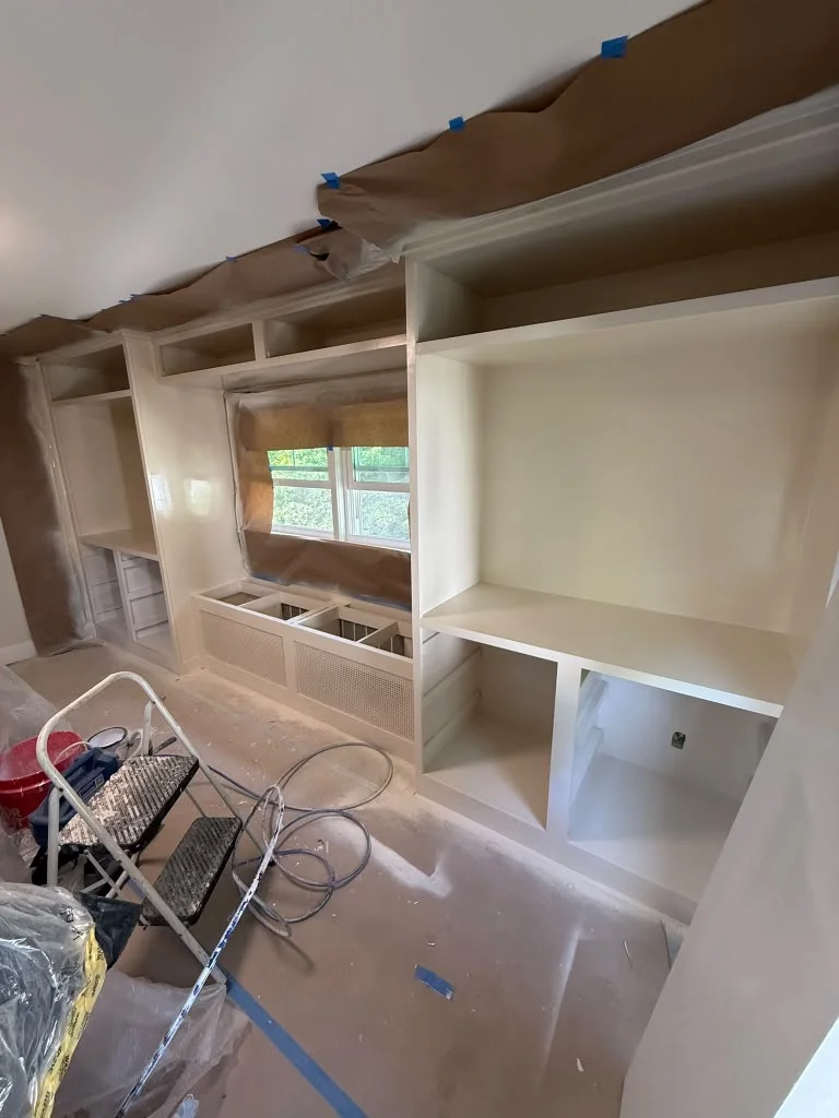 Built-in shelves and cabinetry being painted a light beige color, with drop cloths on the floor. by ALP Home Services in Ever