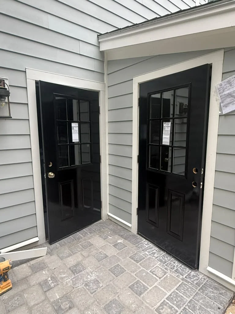 Two freshly painted black exterior doors with white trim on a gray siding wall. by ALP Home Services in Cambridge, MA